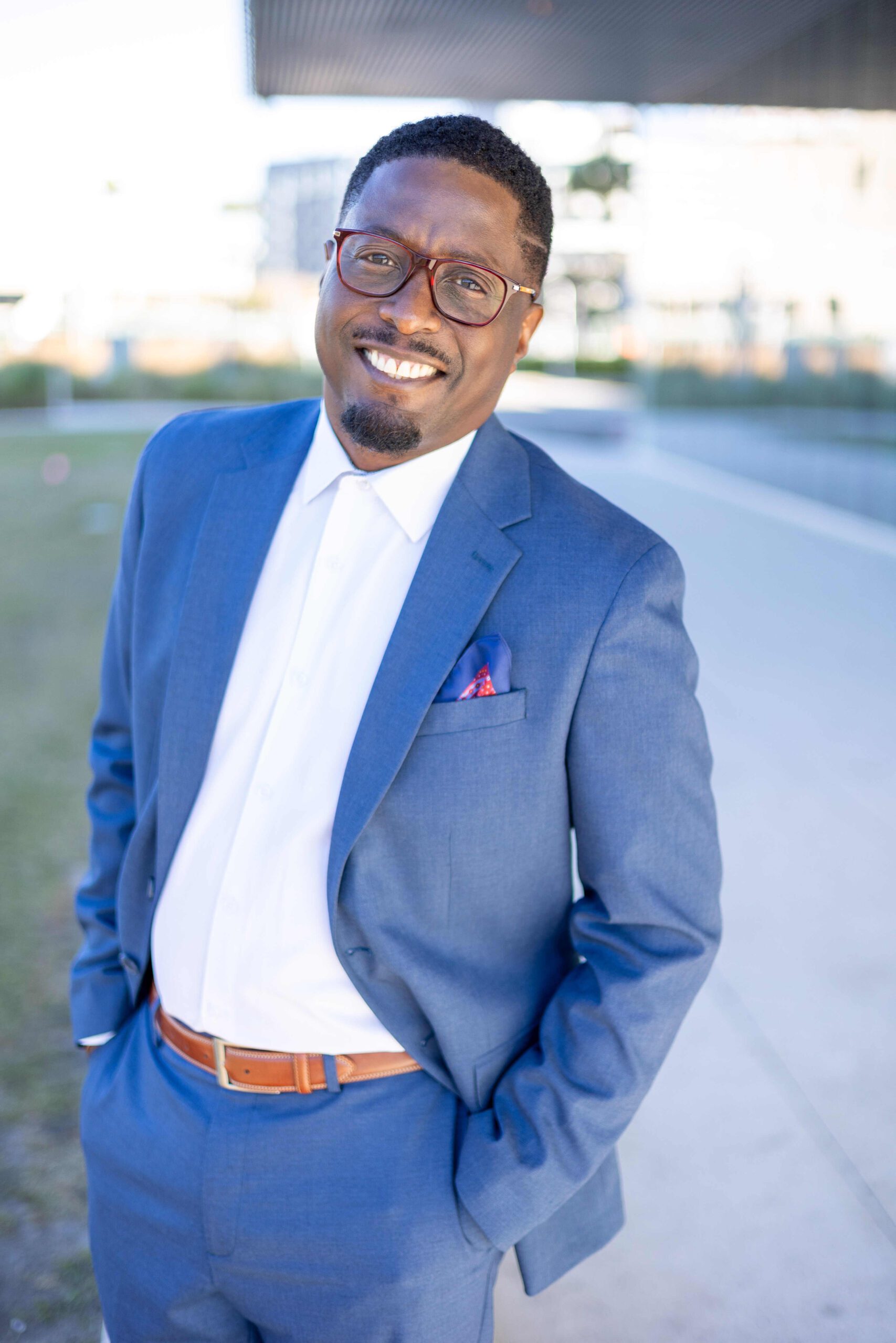 Smiling man in a blue suit posing outdoors with hands in pockets.